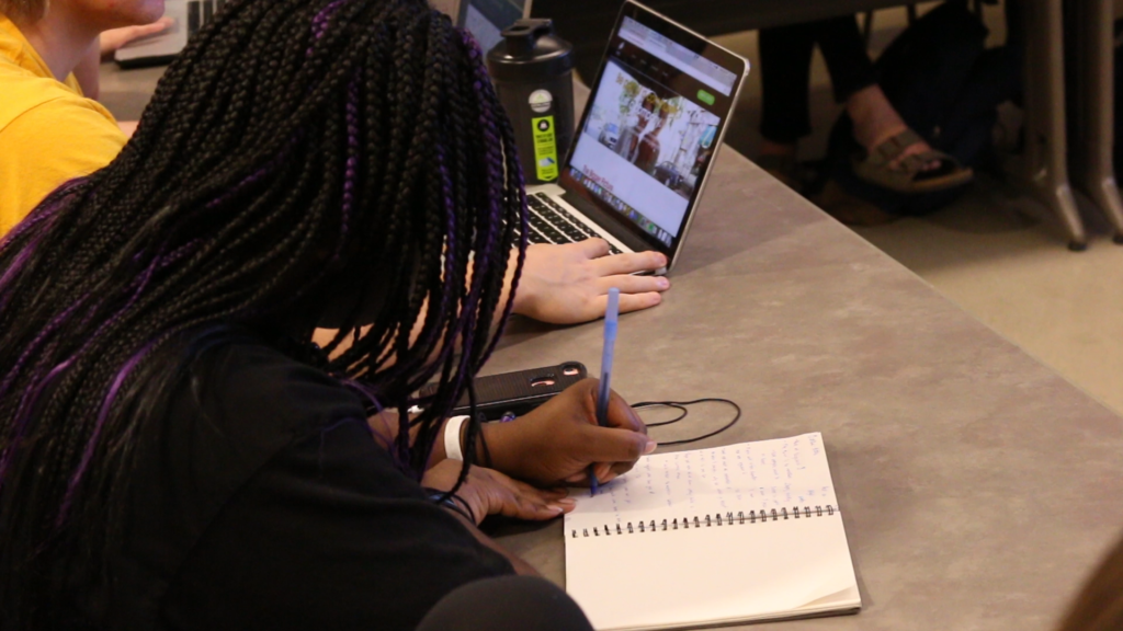 A student in an intermediate screenwriting class writes notes during roundtable feedback on her script outline.