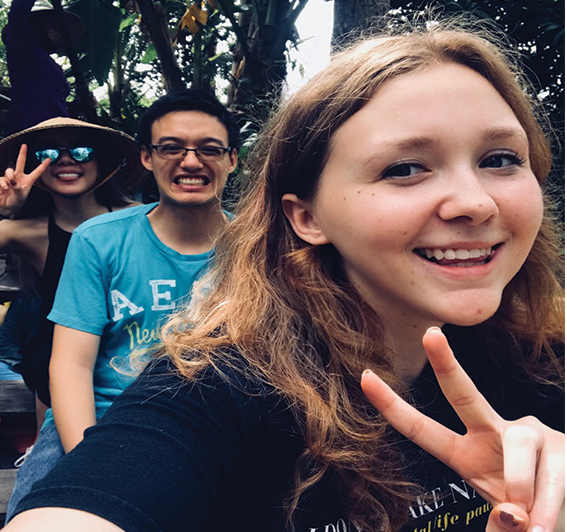 Kaylee Miceli smiles at the camera and gives the peace sign with her fingers as fellow students look on in the background.