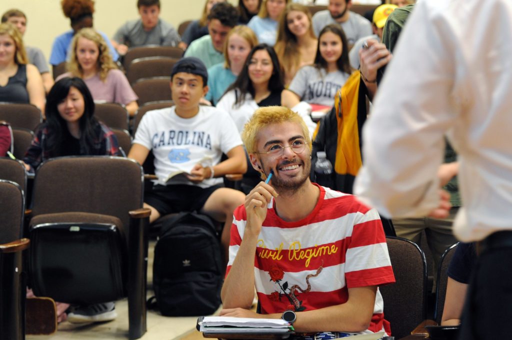 First-year and transfer students can sign up for five new pilot courses in spring 2019 that address broad topics and are team-taught by outstanding faculty members across three different disciplines. Pictured is a large classroom with a student looking toward the professor and other students in the background.