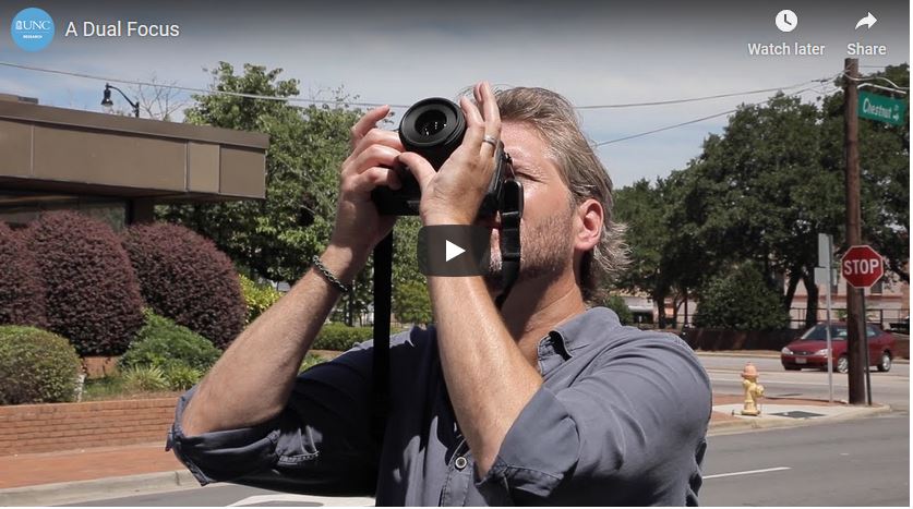 Screen shot shows Ph.D. student John Bechtold holding his camera and looking up at the sky to take a photo.