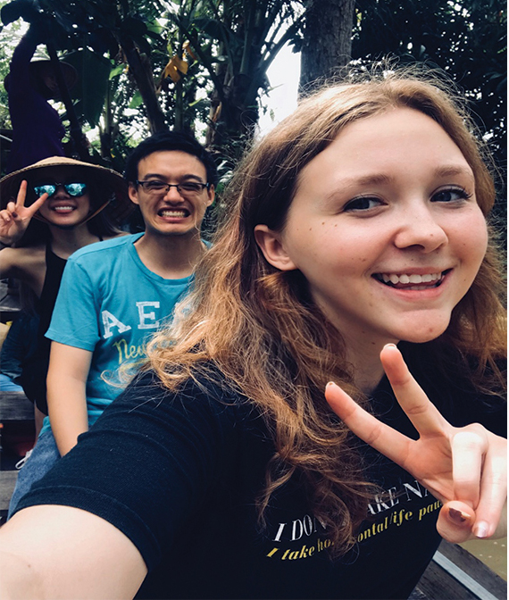 Kaylee Miceli smiles at the camera and gives the peace sign with her fingers as fellow students look on in the background.