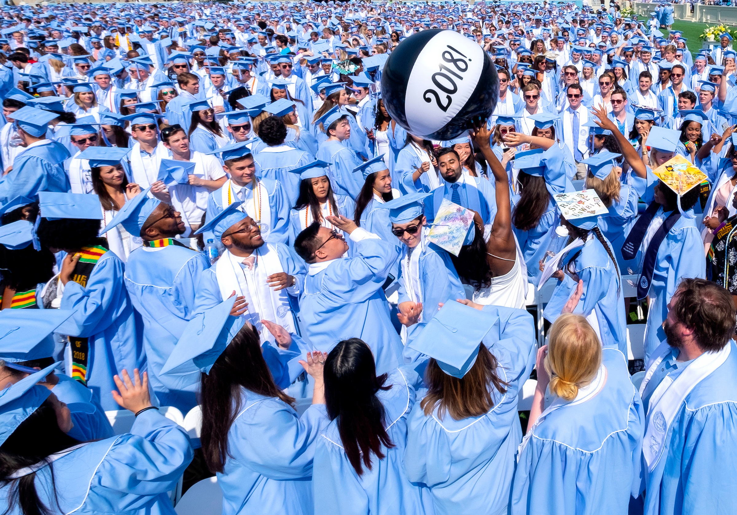 Photo shows the class of 2018 in Carolina blue caps and gowns tossing a black ball in the air that says 2018 on it and the UNC 2018 graduation ceremony.