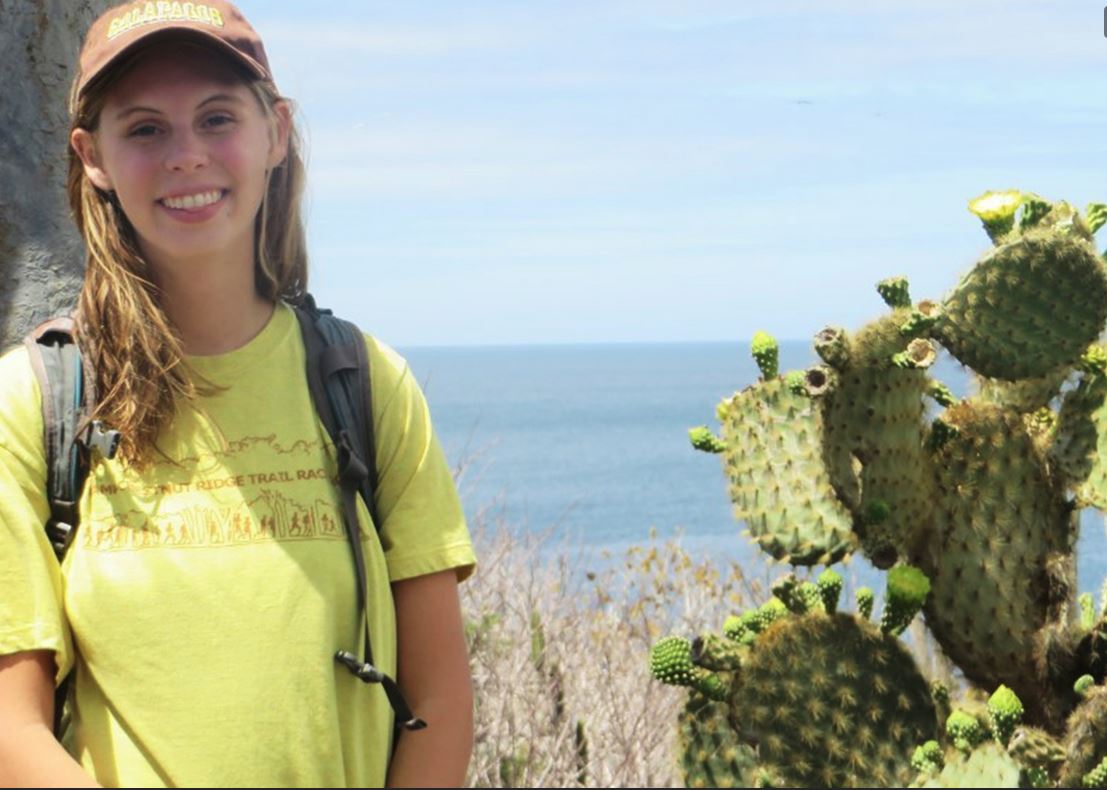 Haley Moser stands in front of a cactus; she is wearing a yellow T-shirt and baseball hat. The baseball hat says "Galapagos" on it. 