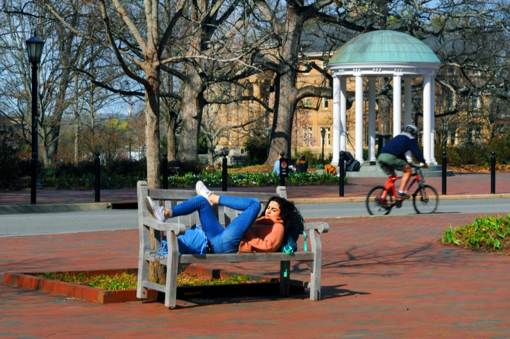 Photo shows a student lying on a bench in front of the Old Well while a biker speeds past on Cameron Avenue. (photo by Donn Young)