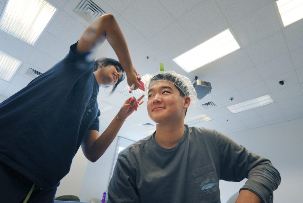 A student sits with a clear plastic-type showercap on his head in the neuroscience class of Marsha Penner. Another student is using a magic marker to make marks on the cap.