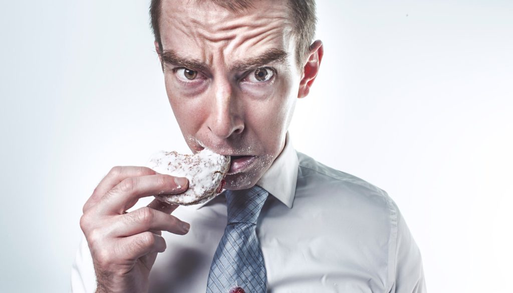 Picture shows a hungry/angry white man wearing a shirt and tie and eating a powdered donut.