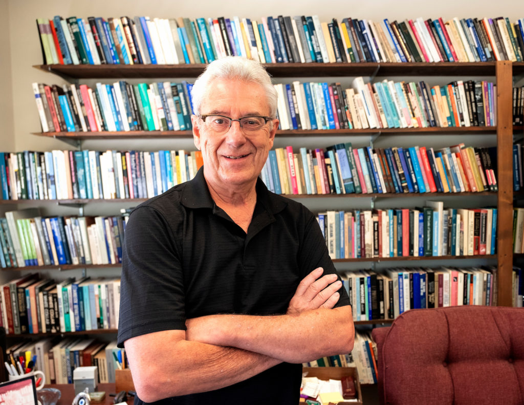 Arne Kalleberg stands in his office with arms crossed, bookshelves filled with books behind him.