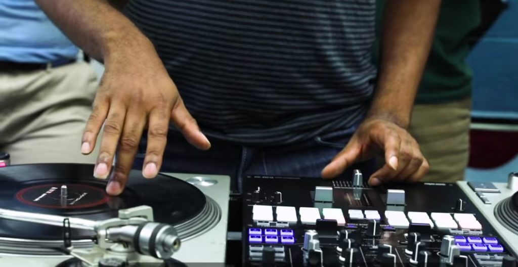 Close up shows a DJ's hands "scractching" a vinyl record on a record player.