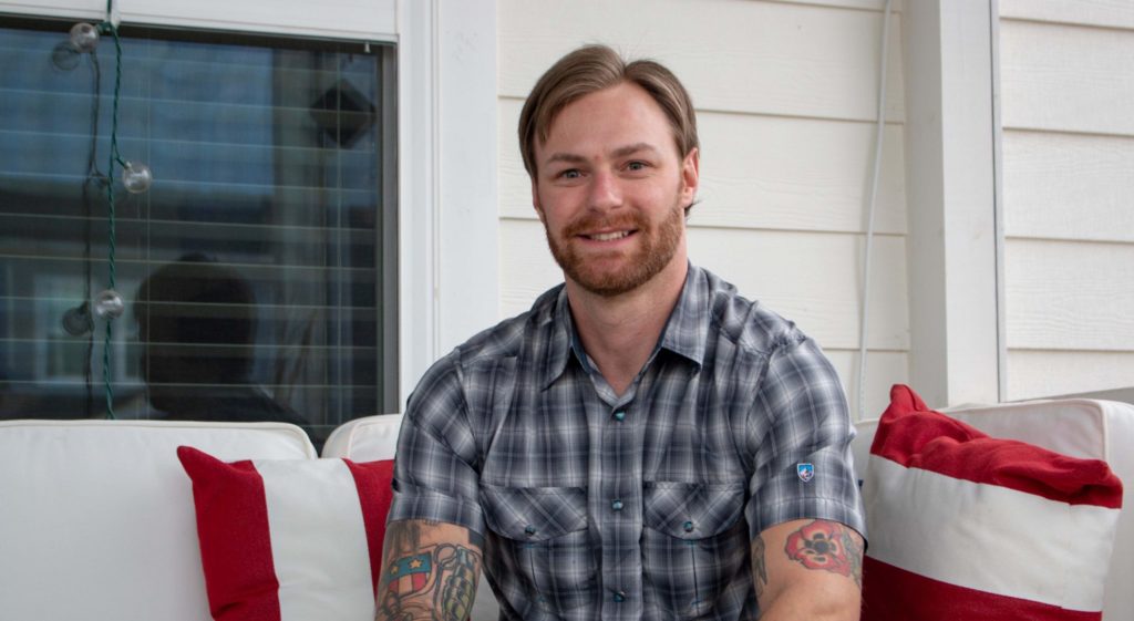 “Don’t be afraid to put yourself out there” is Zac Rhyner’s advice to veterans who want to return to school. He’ll graduate from Carolina in May. (photo by Kristen Chavez). Rhyner is sitting on the front porch of his home with an American flag draped on his sofa seat.