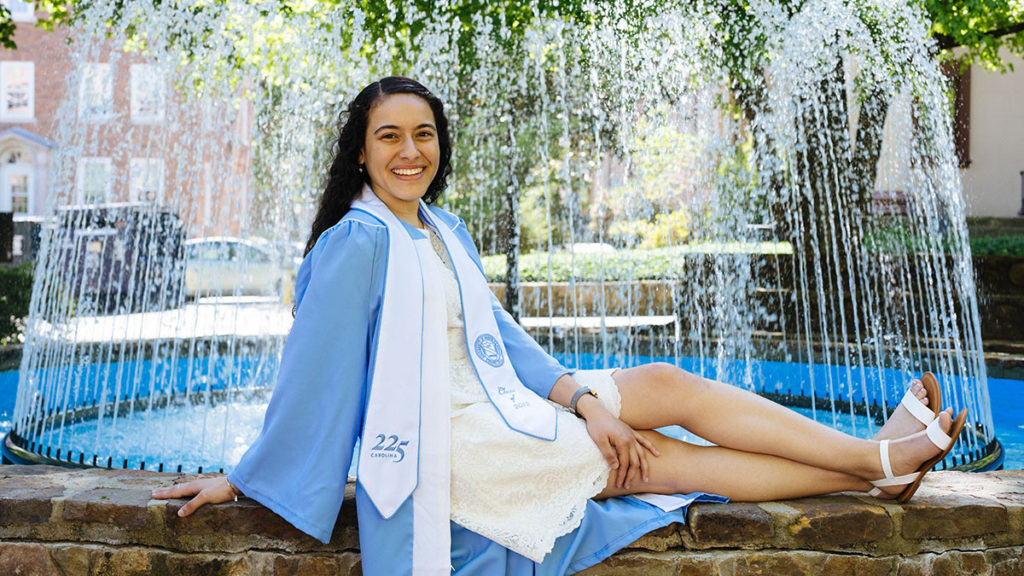 Hanan Alazzam (Photo by Emily Sullivan). She is sitting on the ground in her blue cap and gown and smiling.