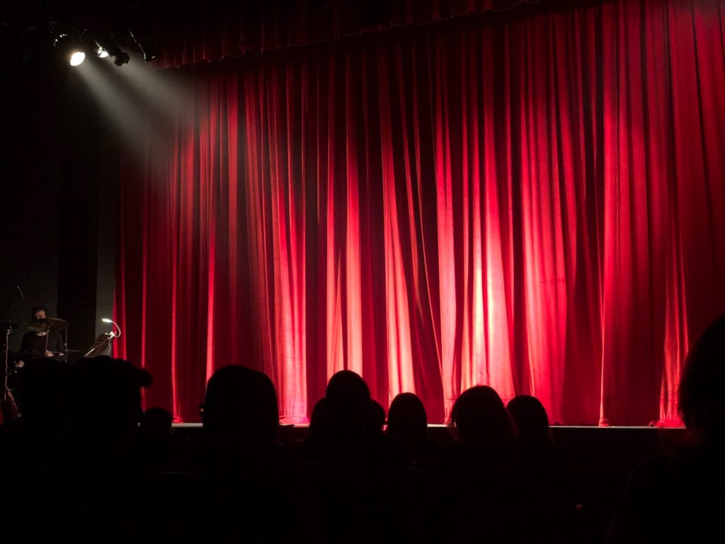 "The Ethics of Stand-up Comedy" examines comedy’s role in society as a form of cultural expression full of underlying ethical discussions.. Photo shows an empty darkened stage with a red curtain and a spotlight and the audience members heads in the foreground.