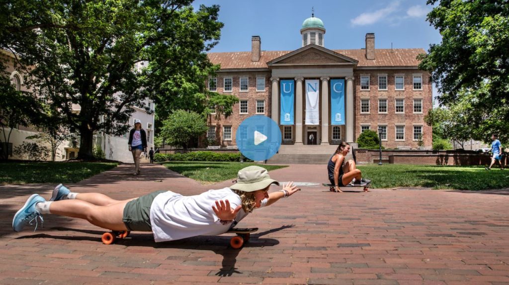 A screenshot of a video showing a girl lying flat on her stomach on a skateboard right in front of South Building on the UNC campus.