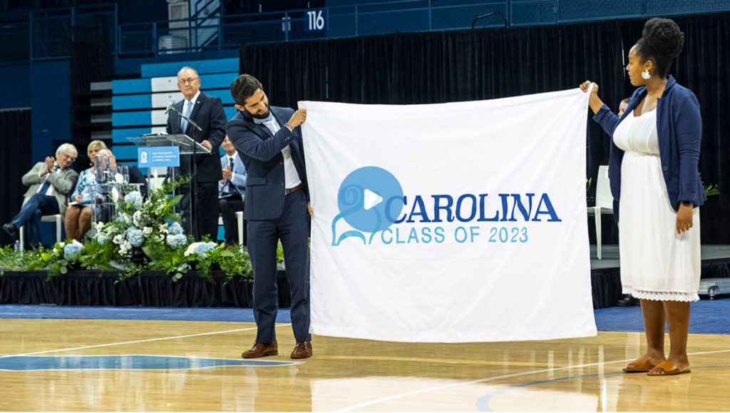 New student convocation pictures shows two people holding up a banner at the event that reads "Carolina Class of 2023".