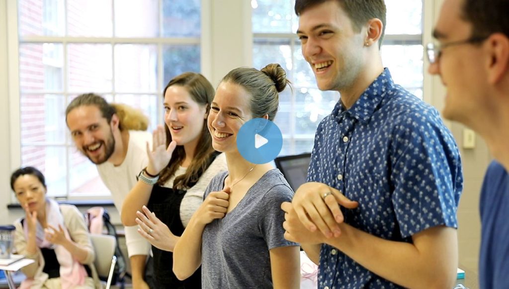 Screen capture shows laughing and smiling students in a classroom doing sign language with their instructor.