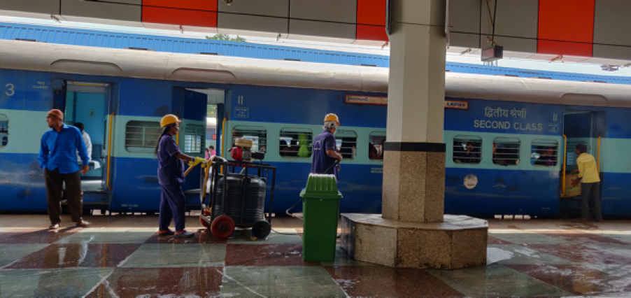 Cleaning staff in blue uniforms with yellow helmets use a jet machine for cleaning the platform and tracks in a railway station.
