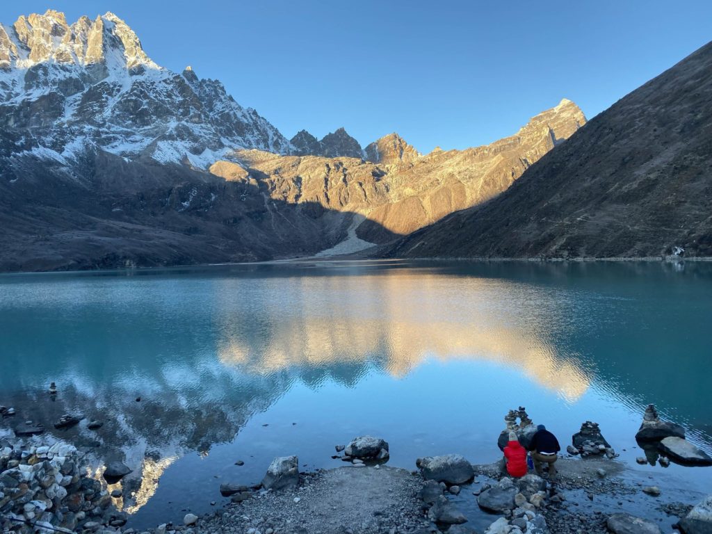Lake 3, early morning. (photo by Roberto Camassa) A crystal blue lake is shown with a lone researcher in a red jacket sitting on the rocks gazing at it with snow covered mountain peaks in background.