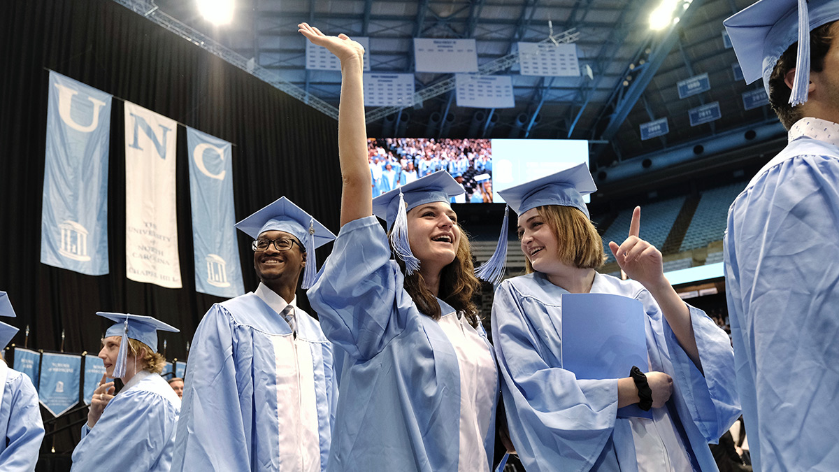 winter graduates in their commencement gowns chat and raise their hands to the ceiling in joy.